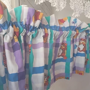 May include: A valance curtain featuring a colorful plaid pattern with Winnie the Pooh and Tigger characters. The curtain has a white background with blue, green, purple, and yellow stripes. The top edge is gathered, and the design is suitable for a child's room.