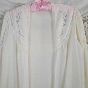 May include: A white, sheer robe with long sleeves, embroidered with small blue and pink flowers. The robe has a lace trim along the neckline and sleeves. It is hanging on a pink satin hanger with a bow.