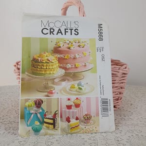 May include: A McCall's Crafts pattern, M5868, featuring images of decorative cakes and cupcakes. The pattern shows a two-tiered pink cake, a ruffled yellow cake, and various cupcake designs. The pattern is in a pink wicker basket.