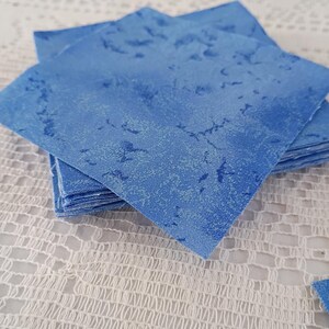 May include: A stack of square fabric pieces in shades of blue. The fabric has a mottled pattern, with darker blue accents on a lighter blue background. The squares are neatly stacked on a white, textured surface.