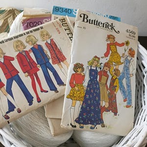 May include: A collection of vintage Butterick sewing patterns featuring illustrations of children's clothing designs. The patterns are in a white basket with spools of white thread. The patterns showcase various outfits, including dresses, overalls, and suits.
