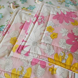 May include: Pink floral quilted potholders with white, yellow, and pink flower designs. The potholders have a white border and a loop for hanging. The background is a floral patterned fabric.