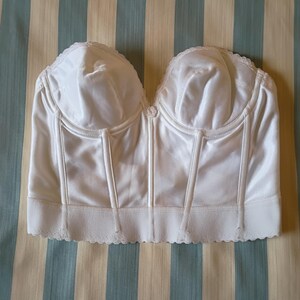 May include: A white satin bustier with a sweetheart neckline and boning details. The bustier has a scalloped lace trim along the bottom edge and a small button detail in the center. The garment is displayed on a striped background.