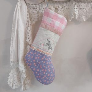 May include: A pink and white gingham fabric Christmas stocking with a purple floral fabric toe and a white fabric panel with an embroidered dragonfly design.