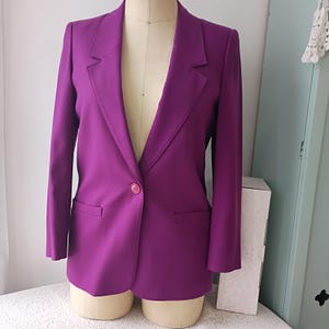 May include: A vibrant purple blazer featuring a single-button closure, notched lapels, and two horizontal welt pockets at the waist. The garment is displayed on a dress form, showcasing long sleeves and a structured, tailored silhouette.