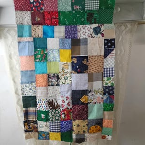 May include: A patchwork quilt made of various fabric squares in a grid pattern. The squares feature a variety of colors and patterns, including floral, plaid, and solid colors. The quilt is hanging, with a white lace curtain visible on the side.
