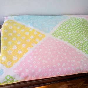 May include: A folded sheet featuring a patchwork design with pastel colors. The sheet has four sections: yellow, pink, green, and blue, each with a scalloped edge and a white floral pattern. The sheet is made of a soft fabric.