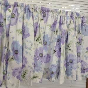 May include: A floral valance with a white background and large purple and light blue flowers. Green leaves and smaller blue flowers are also present. The valance is gathered at the top and hangs in soft folds.