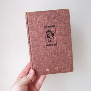 May include: A brown hardcover book with a black and white illustration of a woman wearing a nurse's uniform and a cap. The book's title is "The Nurse"