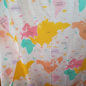 May include: A white fabric with a colorful world map design. The map features continents in shades of yellow, pink, orange, and green, with animal illustrations and country names. The grid lines and text are in black.