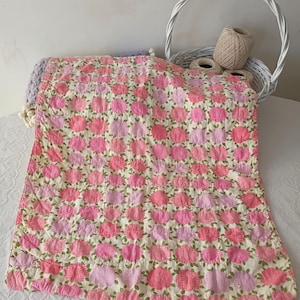 May include: A handmade quilt featuring a floral pattern in shades of pink and light pink on a cream background. The quilt is adorned with green leaves and is likely made of cotton. A white basket with yarn is in the background.