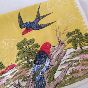 May include: A yellow linen napkin featuring a vintage bird print. The design includes two birds with red heads, blue and black wings, and a landscape with trees and a red barn. The napkin has a white, scalloped edge.