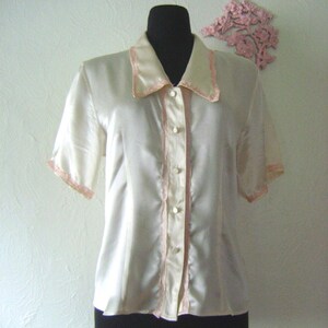 May include: Cream-colored short-sleeve blouse with a collared neckline and button-down front. The blouse features pink trim along the collar, button placket, and sleeve edges. The garment is displayed on a black mannequin.