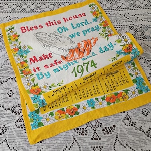 May include: Vintage 1974 kitchen towel with floral border and calendar. The towel features the text "Bless this house, Oh Lord, we pray, Make it safe by night and day." The calendar shows the months of January, February, March, and December.
