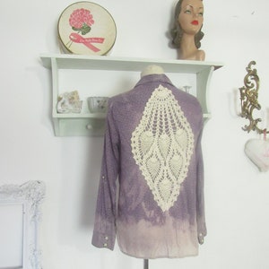 May include: A purple button-down shirt with a cream-colored, openwork crochet design on the back. The shirt has long sleeves and pearl-like buttons. The bottom of the shirt fades to a lighter shade of purple.