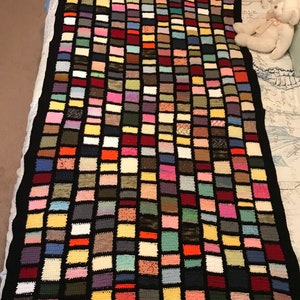 May include: A colorful crocheted blanket with a black border. The blanket is made up of small squares of different colors, including red, blue, green, yellow, and purple.