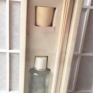 May include: A white box with a clear plastic window showing a glass bottle of diffuser oil, a white ceramic diffuser base, and a bundle of wooden diffuser sticks.