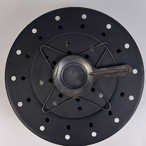 May include: A circular black metal stove top heat diffuser plate featuring a series of evenly spaced circular perforations. Centered on the plate is a four-pronged wire support rack holding a small, round, dark grey metal pan with a handle.