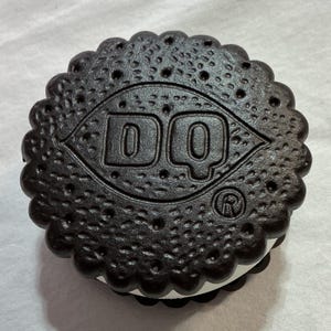 May include: A round, dark brown sandwich cookie with a scalloped edge and a textured surface. The center features the embossed letters DQ inside a diamond shape, with a registered trademark symbol. A white cream filling is visible between the two wafers.