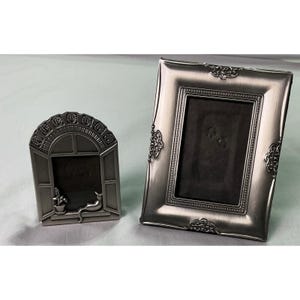 Puede incluir: Dos marcos de fotos de metal en tono plateado. Un marco es rectangular con detalles de filigrana en los bordes. El segundo marco es arqueado, con forma de ventana, que incluye una pequeña figura de gato en el alféizar y una planta.