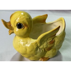 May include: A glossy yellow ceramic duck planter featuring a bright yellow body, a smooth orange beak, and dark eyes. The duck has sculpted wing details on its sides and a hollow interior space, designed with a smooth, reflective glazed finish.