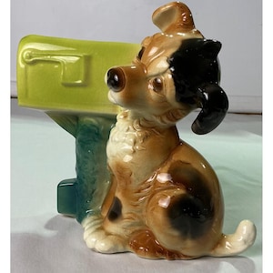 May include: A ceramic figurine of a seated dog with brown, black, and white fur, positioned next to a lime green mailbox mounted on a textured, dark teal post. The dog has a dark brown nose and ears, with a glossy finish throughout the piece.