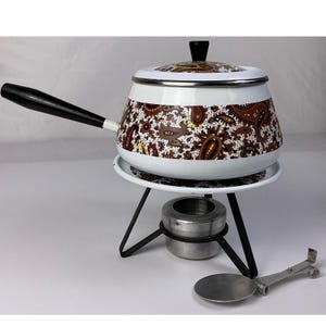 May include: A white enamel fondue pot featuring a brown and black paisley pattern, a black handle, and a matching lid with a black knob. It sits on a black metal stand with a silver burner and includes a separate silver metal tool with a flat end.