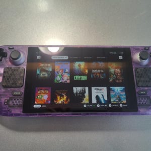 May include: A purple handheld gaming console with a screen displaying a variety of video game titles. The console has two buttons on the top and two on the bottom, as well as a directional pad and four action buttons on the right side. The screen shows a variety of games, including "Deadlight", "Cryptark", and "Dying Light 2".