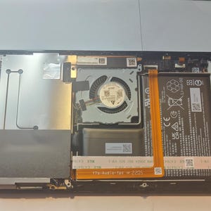 May include: Close-up view of the inside of a disassembled gaming console. The motherboard is visible, along with the cooling fan, battery, and other components. The console is black and has a silver heat sink.