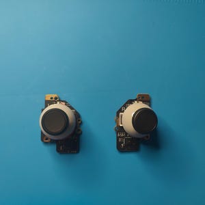 May include: Two black and white analog joystick controllers with circuit boards. The joysticks are on a blue surface.