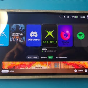 May include: A clear handheld gaming device with a black screen displaying a variety of app icons, including Discord, XEMU, Firefox, and Spotify. The device has a black and white button layout and a black joystick.
