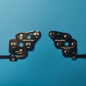 May include: Two black circuit boards with buttons and the text "eXtremeRate" are displayed against a blue background. One board has "LEFT" and "RIGHT" labels, while the other has "Menu". The boards are components for a gaming device.