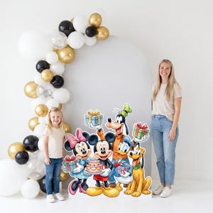 May include: A large, colorful cardboard cutout featuring Mickey Mouse, Minnie Mouse, Donald Duck, Goofy, and Pluto holding birthday gifts and a cake. The display is positioned next to a balloon arch made of white, black, and metallic gold balloons.