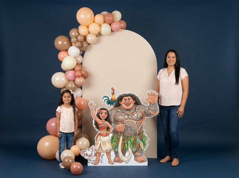May include: A party scene featuring a large cardboard cutout of characters from the animated film Moana, including Maui, Moana, and Hei Hei. A balloon arch in shades of beige, pink, and brown frames a beige backdrop. A woman and a young girl stand nearby.