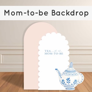 Peut inclure: Un fond de scène rose clair en forme d'arche avec un bord festonné, avec le texte "TEA with the MOM-TO-BE". Une théière décorative avec des motifs floraux bleus est à droite. Le fond est posé contre un mur clair et un sol en bois.