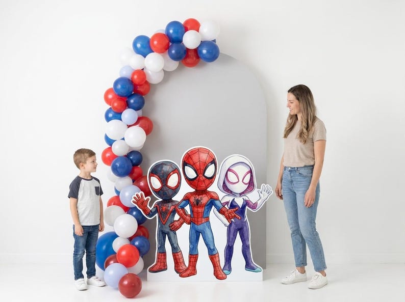 May include: A party scene featuring a balloon arch in red, white, and blue, with three Spider-Man character cutouts. A young boy and a woman stand nearby. The scene is set against a white backdrop.