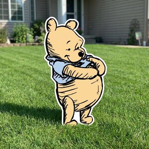 May include: A flat, cutout-style yard sign featuring a cartoon bear with light yellow fur and a light blue shirt. The bear is depicted in a standing position with its arms crossed over its round belly, featuring black outlines and shading.