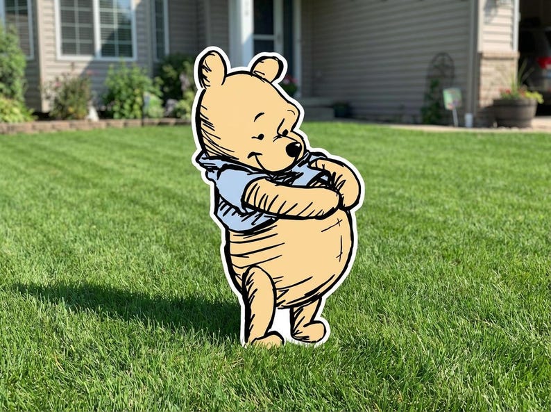 May include: A flat, cutout-style yard sign featuring a cartoon bear with light yellow fur and a light blue shirt. The bear is depicted in a standing position with its arms crossed over its round belly, featuring black outlines and shading.