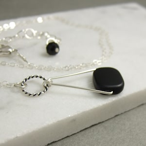 May include: A silver necklace featuring a pendant with a black, rectangular stone. The necklace has a delicate chain and a twisted silver ring detail. The pendant is attached with a silver pin-like design.