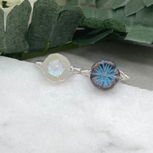 May include: Two silver wire wrapped rings with glass flower accents. One ring has a white iridescent flower and the other has a dark brown flower with a blue center.