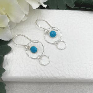 May include: Silver dangle earrings with a turquoise blue gemstone in the center of a textured circle, and a smaller textured circle dangling below.
