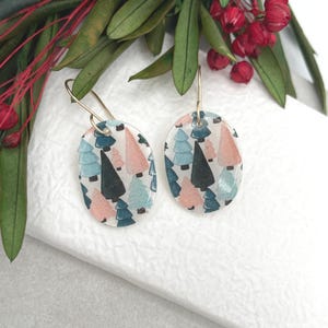 May include: Oval-shaped earrings with a festive Christmas tree pattern in pink, teal, and black on a white background. The earrings have gold-colored ear wires and are displayed with greenery and red berries.