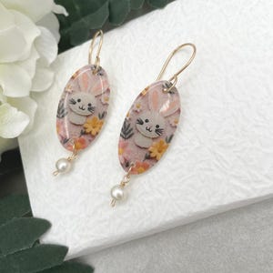 Pode incluir: Um par de brincos de argola dourados com pingentes de resina ovais que apresentam um coelho branco com orelhas cor-de-rosa e um padrão floral. Cada pingente tem uma única pérola branca pendurada na parte inferior.
