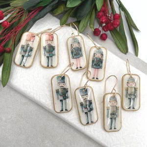 May include: Eight pairs of gold-tone dangle earrings with rectangular resin charms featuring vintage-inspired illustrations of nutcracker soldiers in blue, red and white.