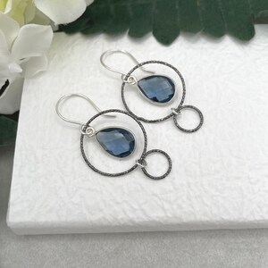 May include: A pair of silver earrings with a dark blue teardrop-shaped gemstone hanging from a black wire circle. Each earring has a smaller black wire circle dangling below the larger circle.