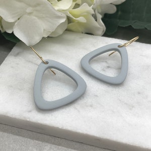 May include: A pair of gray triangle shaped earrings with gold hooks. The earrings are made of a matte material and have a simple, modern design.