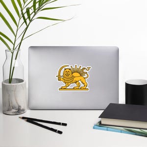 Op de afbeelding: Een sticker met een gele leeuw met een zwaard en een zonnestraalontwerp erachter. De leeuw staat op een gele rechthoekige basis. De sticker zit op een grijze laptop.