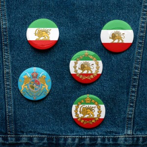 May include: Five circular pin badges showcasing the Iranian flag and emblem. The badges feature a green, white, and red colour scheme, with a gold lion and sun emblem. The badges are displayed on a denim background.