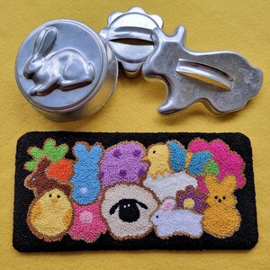 Puede incluir: Una alfombra de fieltro negro con un diseño bordado de colores de varios animales con temática de Pascua, incluyendo un cordero, pollitos y conejos. La alfombra está rodeada de cortadores de galletas de metal con forma de conejo, flor y pájaro.