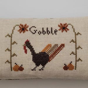 May include: A rectangular decorative pillow with a beige center featuring a cross-stitched turkey design with the word "Gobble". The pillow has green and brown checkered fabric on the sides.